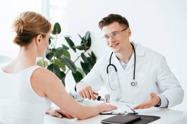 Neşeli dermatolog hastaya bakarken elinde dermatoskop ve gülümsüyor. 