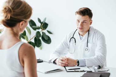 beyaz önlüklü doktorun seçici odak sıkıştırılmış elleri ile oturan ve hasta bakarak 