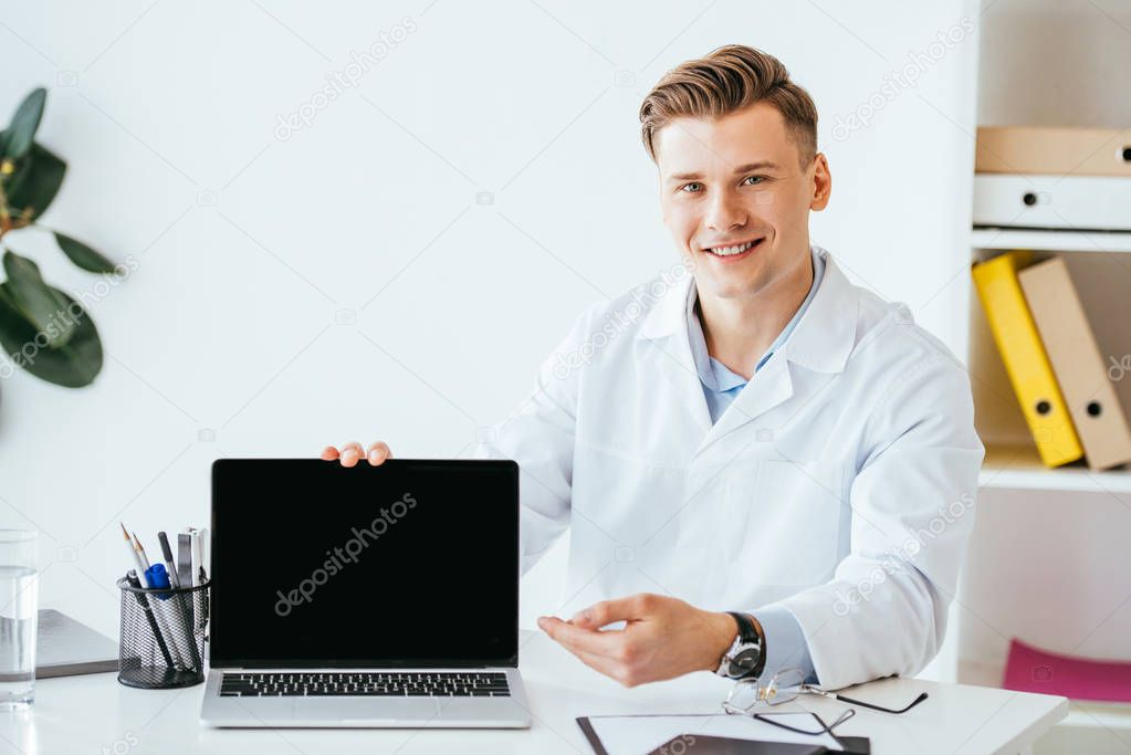 cheerful doctor in white coat sitting near laptop with blank screen 