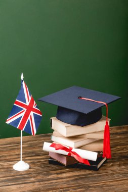 Yeşil ahşap yüzeyüzerinde kitap, akademik şapka, diploma ve İngiliz bayrağı
