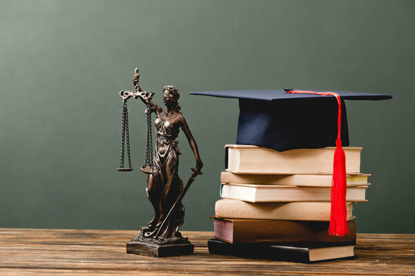 stack of books, themis statuette and academic cap on wooden surface on grey