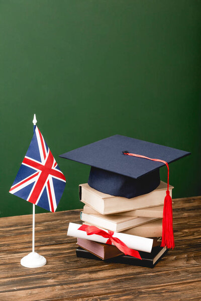 Books, academic cap, diploma and british flag on wooden surface on green