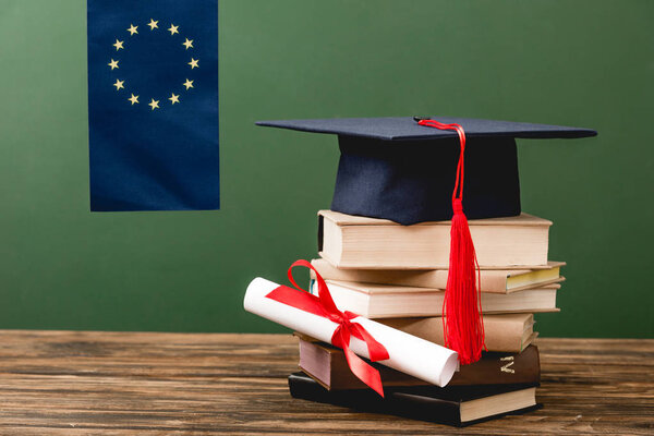 european flag, stack of books, diploma and academic cap on wooden surface isolated on green