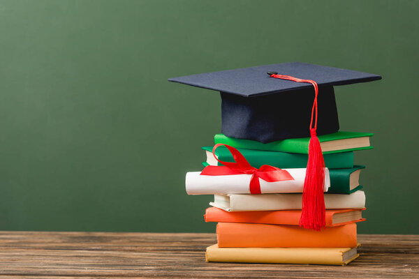 books, diploma with ribbon and academic cap on wooden surface isolated on green