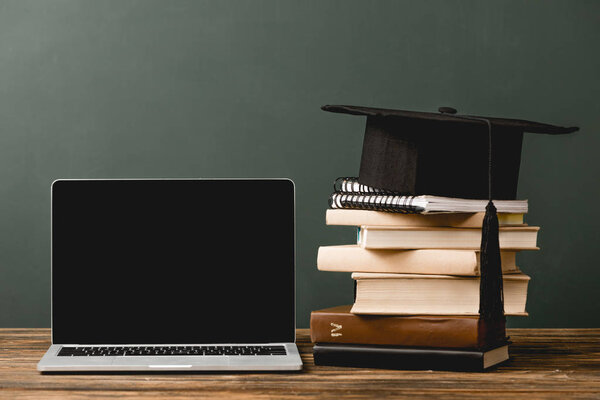 books, notebooks, academic cap and laptop with blank screen on wooden surface isolated on grey