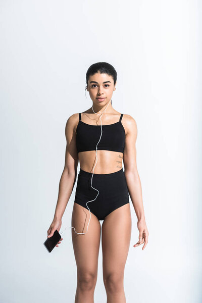 attractive sportive african american girl holding smartphone and listening music in earphones on white