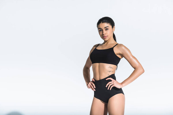 beautiful sportive african american girl holding hands on hips and looking at camera on white