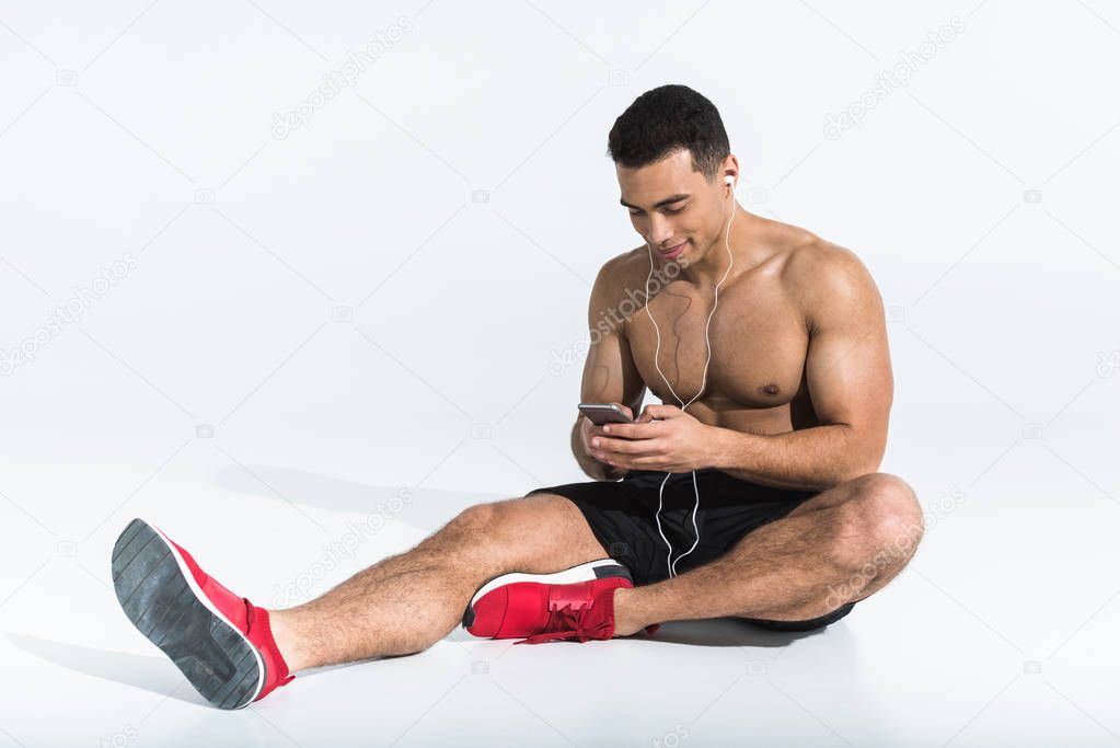 handsome sportive mixed race man sitting on floor and listening music in earphones on white