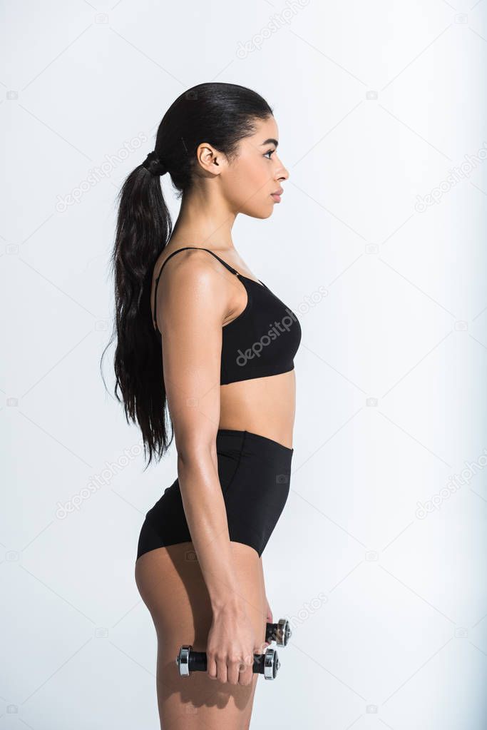 side view of pretty sportive african american girl in black sportstwear holding dumbbells on white