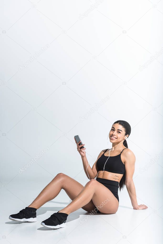 smiling sportive african american girl sitting on floor and listening music in earphones on white