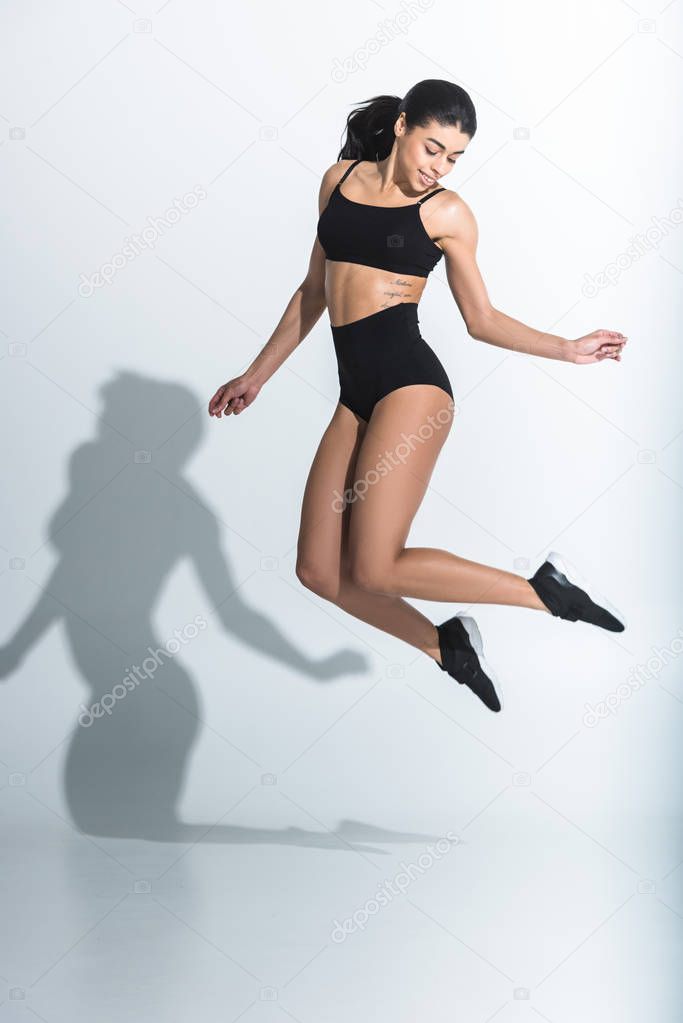 beautiful sportive african american girl in black sports bra and shorts jumping on white