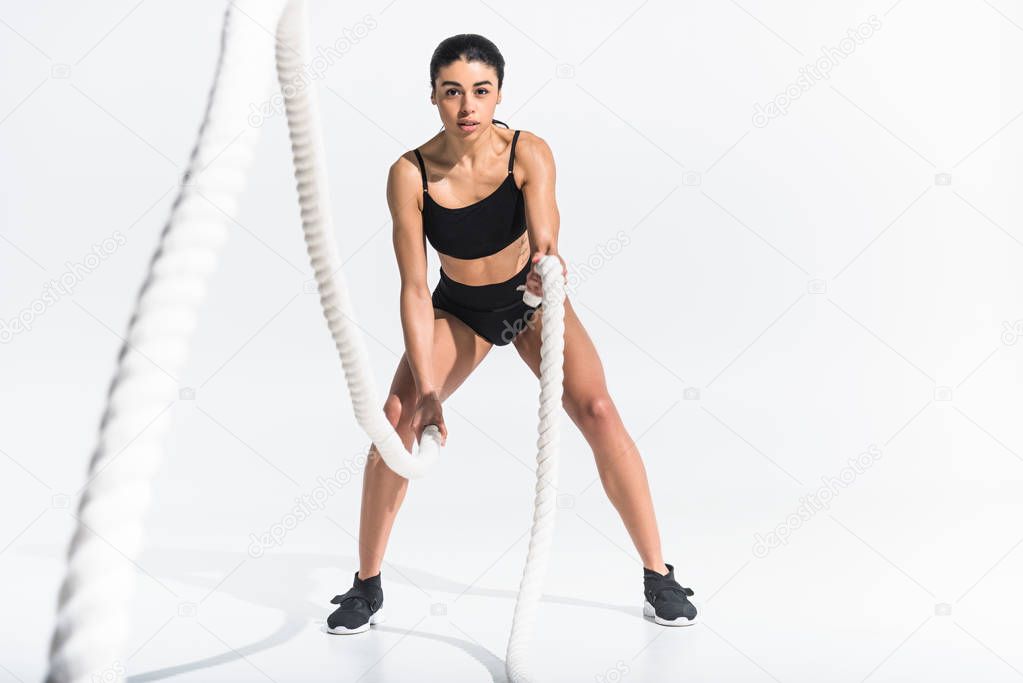 selective focus of pretty sportive african american girl training with white ropes and looking at camera on white