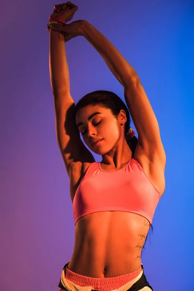 pretty african american girl in ink sports bra stretching on purple and blue gradient background