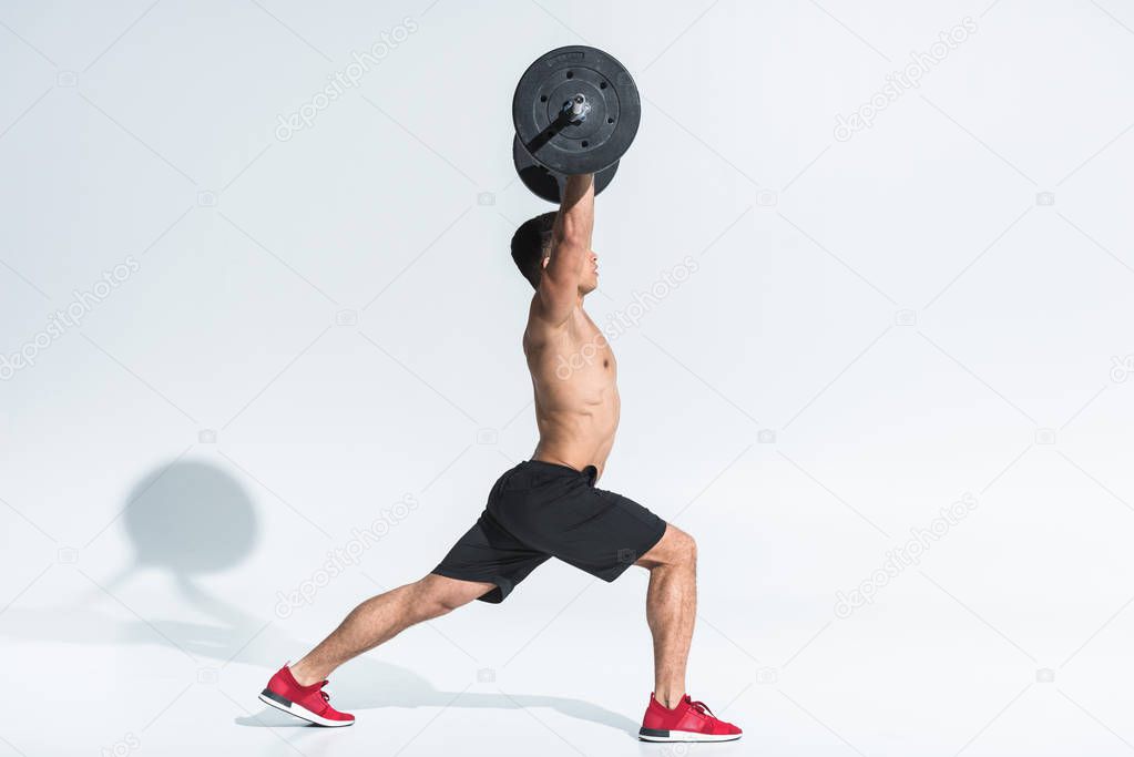 side view of athletic mixed race man in black shorts and red sneakers lifting barbell on white 