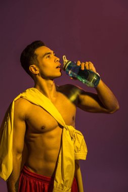 handsome sportive mixed race man drinking from sport bottle on purple background clipart