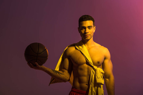 handsome sportive mixed race man holding brown ball on purple background