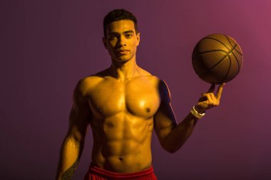 good-looking sportive mixed race man holding brown ball and looking at camera on purple background clipart
