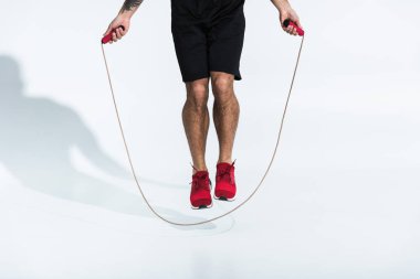 partial view of man in black shorts and red sneakers jumping with skipping rope on white clipart