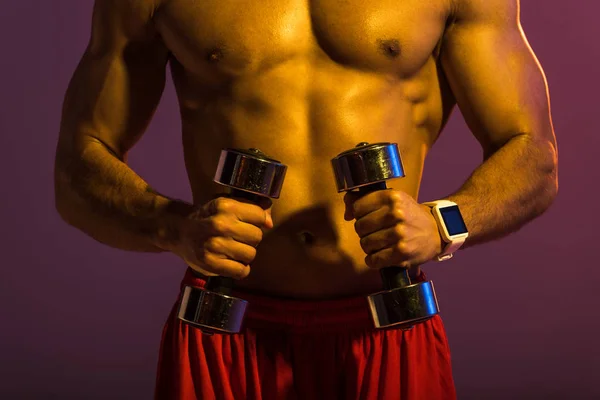 cropped view of shirtless sportive man with dumbbells on purple background