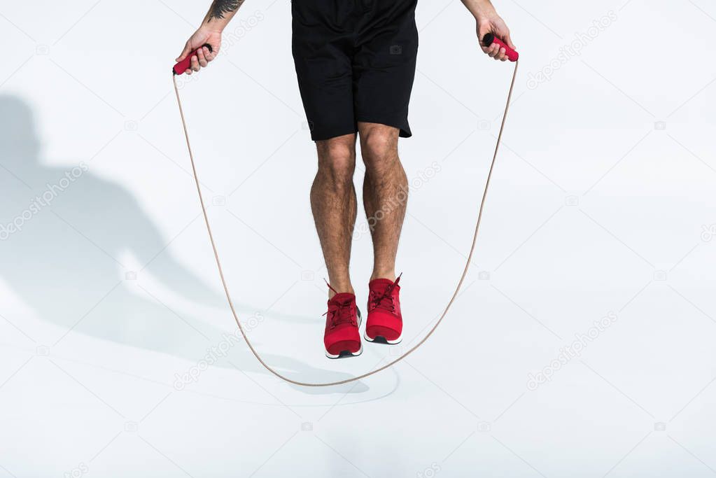 partial view of man in black shorts and red sneakers jumping with skipping rope on white