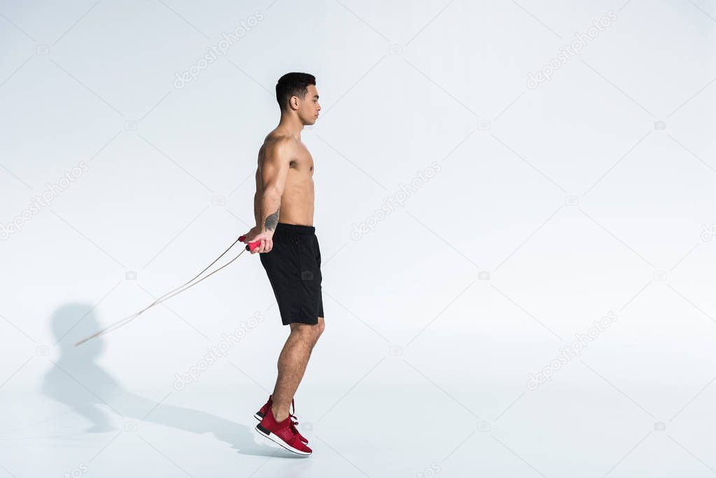 side view of sportive mixed race man in black shorts and red sneakers jumping with skipping rope on white