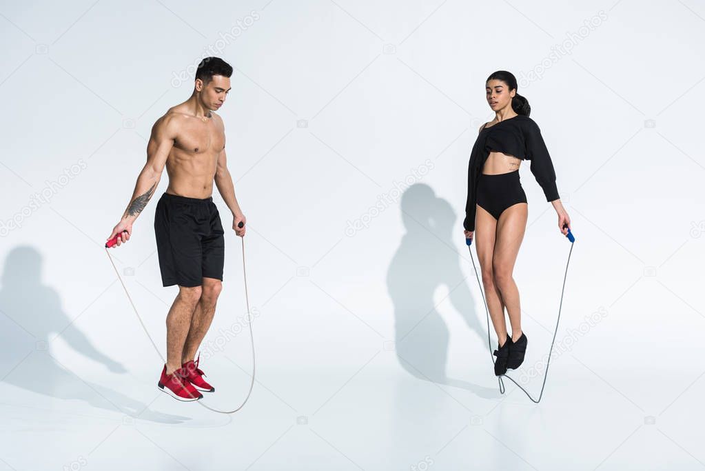 athletic mixed race man and sportive african american woman jumping with skipping ropes on white