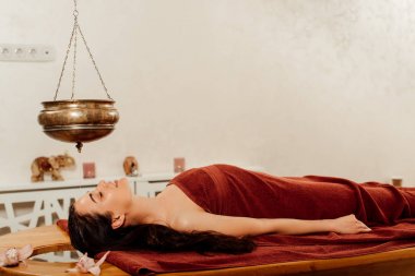 side view of woman lying under shirodhara vessel during ayurvedic procedure clipart