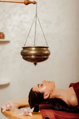 side view of woman lying under shirodhara vessel during ayurvedic procedure clipart