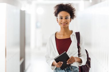 front view of african american student with notebook in corridor in university clipart