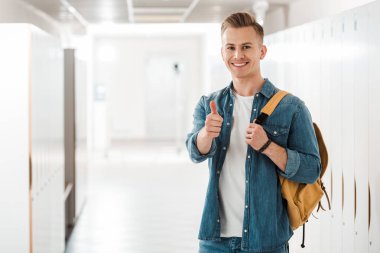 student with backpack showing thumb up in corridor in university clipart