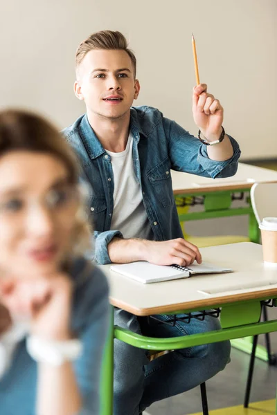 机に座って鉛筆で手を上げるデニムシャツの学生 — ストック写真