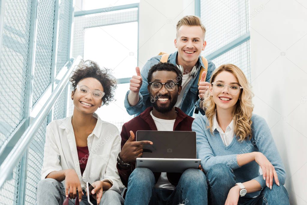 group of smiling multicultural students with laptop showing thumbs up in university