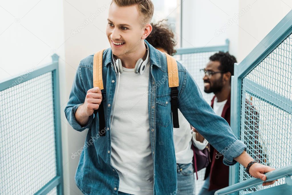 smiling multiethnic students with backpacks on stairs in college