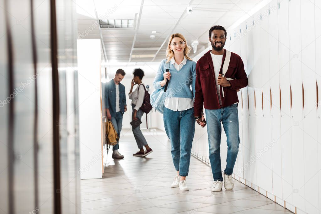 full length view of smiling multiethnic students holding hands in college