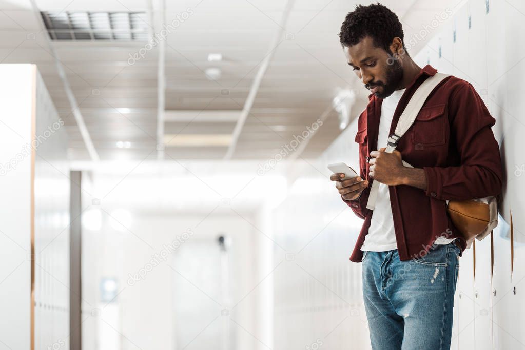 african american student with backpack using smartphone in corridor