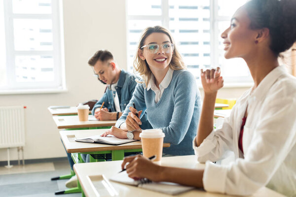 smiling multicultural students with notebooks talking in college