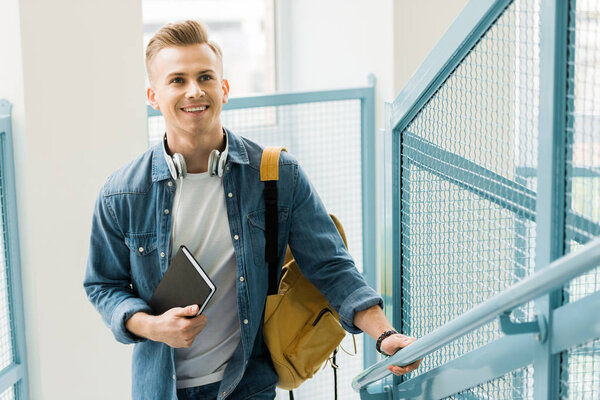 улыбающийся студент в джинсовой рубашке с желтым рюкзаком и блокнотом в университете
