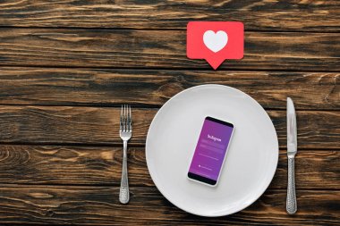 kahverengi ahşap yüzeyüzerinde kalp sembolü ile bıçak, çatal ve kırmızı kağıt kesme kartı yakınında ekranda instagram uygulaması ile akıllı telefon üst görünümü
