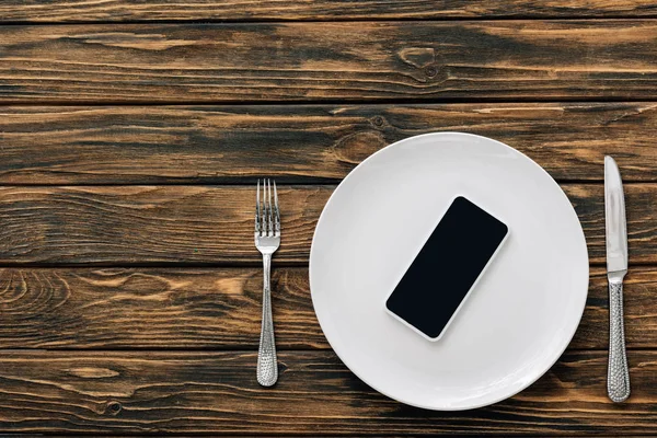 茶色の木製の表面にナイフとフォークの近くの白いプレート上の空白の画面を持つスマートフォンのトップビュー — ストック写真