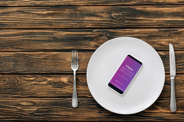 top view of smartphone with instagram app on screen on white plate near knife and fork on brown wooden surface