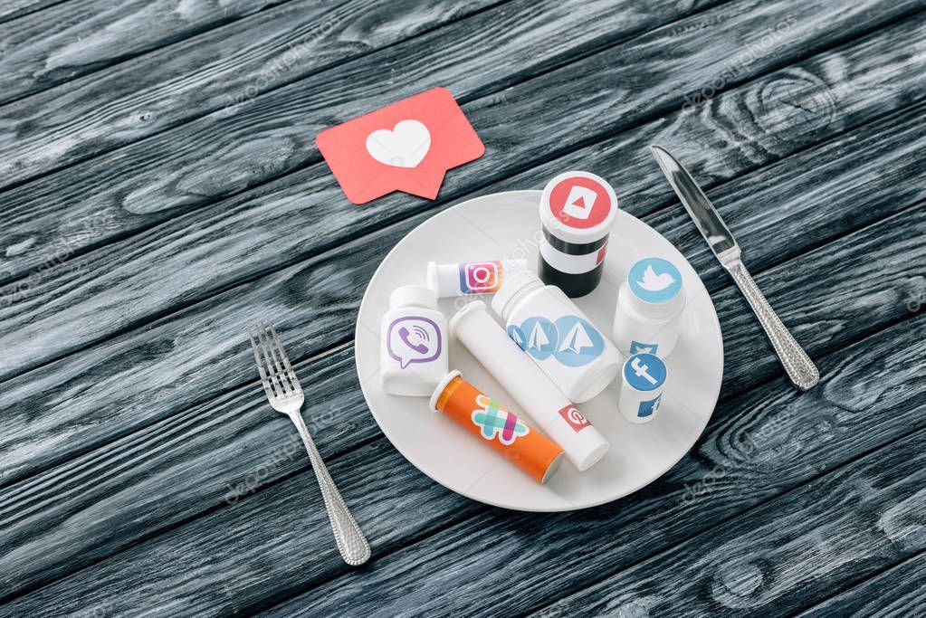 Containers with social media logos on white plate near red paper cut card with heart symbol, knife and fork on grey wooden surface