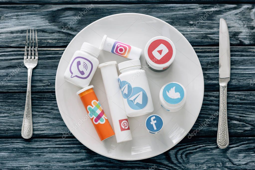 Top view of container with social media logos on white plate near knife and fork on grey wooden surface