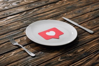 kahverengi ahşap yüzeyüzerinde bıçak ve çatal yakınında beyaz plaka üzerinde kalp sembolü ile kırmızı kağıt kesme kartı