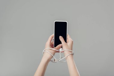 partial view of young woman with usb cable around hands holding smartphone with blank screen isolated on grey clipart