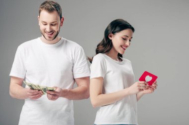 attractive girl holding red paper cut card with heart symbol near handsome man counting dollar banknotes isolated on grey clipart