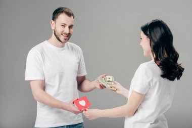 gri arka plan üzerinde kalp sembolü ile kırmızı kağıt kesme kartı ile genç kadına dolar banknotlar veren gülümseyen yakışıklı adam