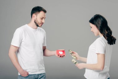 Gri arkaplanda kalp sembolü olan kırmızı kağıt kesim kartı olan yakışıklı bir adama dolar banknotları veren genç bir kadın.