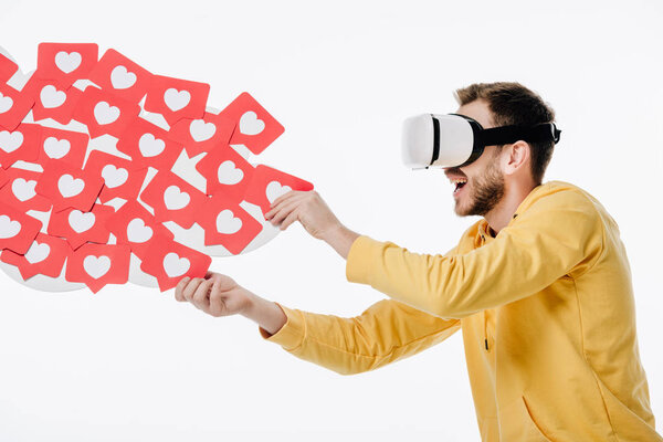 excited man using virtual reality headset while holding red paper cut cards with hearts symbols isolated on white
