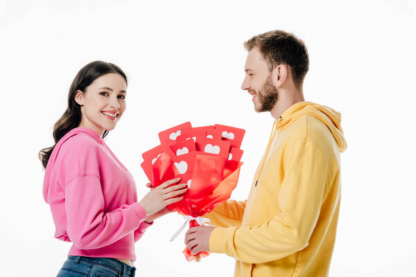 happy young girl taking bouquet of red paper cut cards with hearts symbols from handsome boyfriend isolated on white