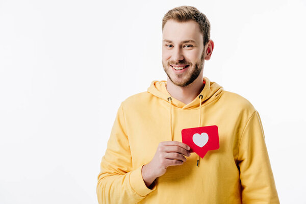 handsome cheerful man in yellow hoodie holding red paper cut card with heart symbol isolated on white